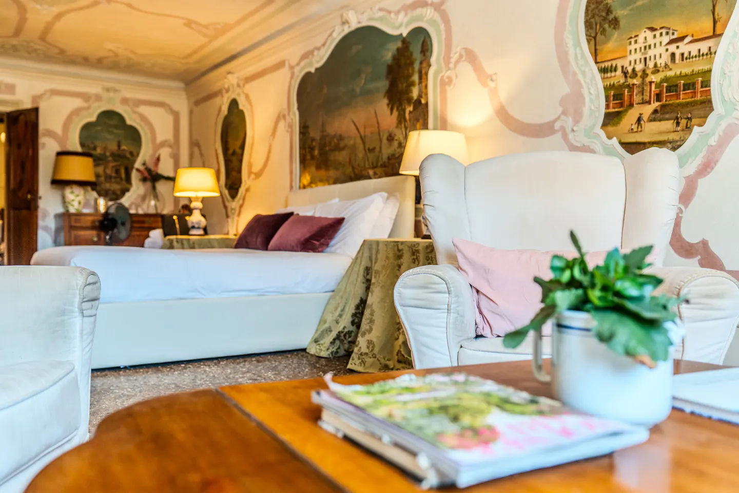Elegant bedroom with white armchairs, a bed with purple pillows, ornate wall murals, and a wooden table with a plant.