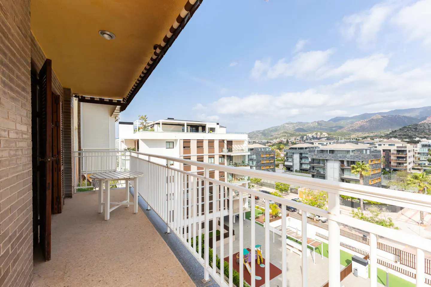 Balcony view with white railings overlooking a courtyard with playground equipment, buildings, and mountains in the distance.