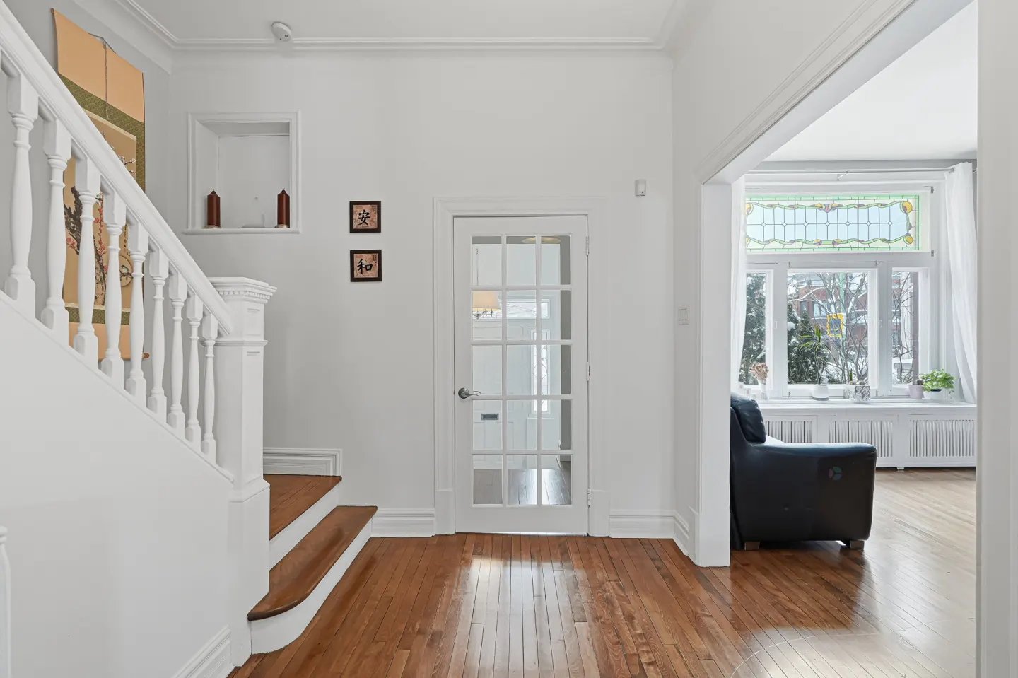 Bright foyer with hardwood floors, white walls, and staircase. A glass-paneled door and an open doorway lead to a living room with a window.