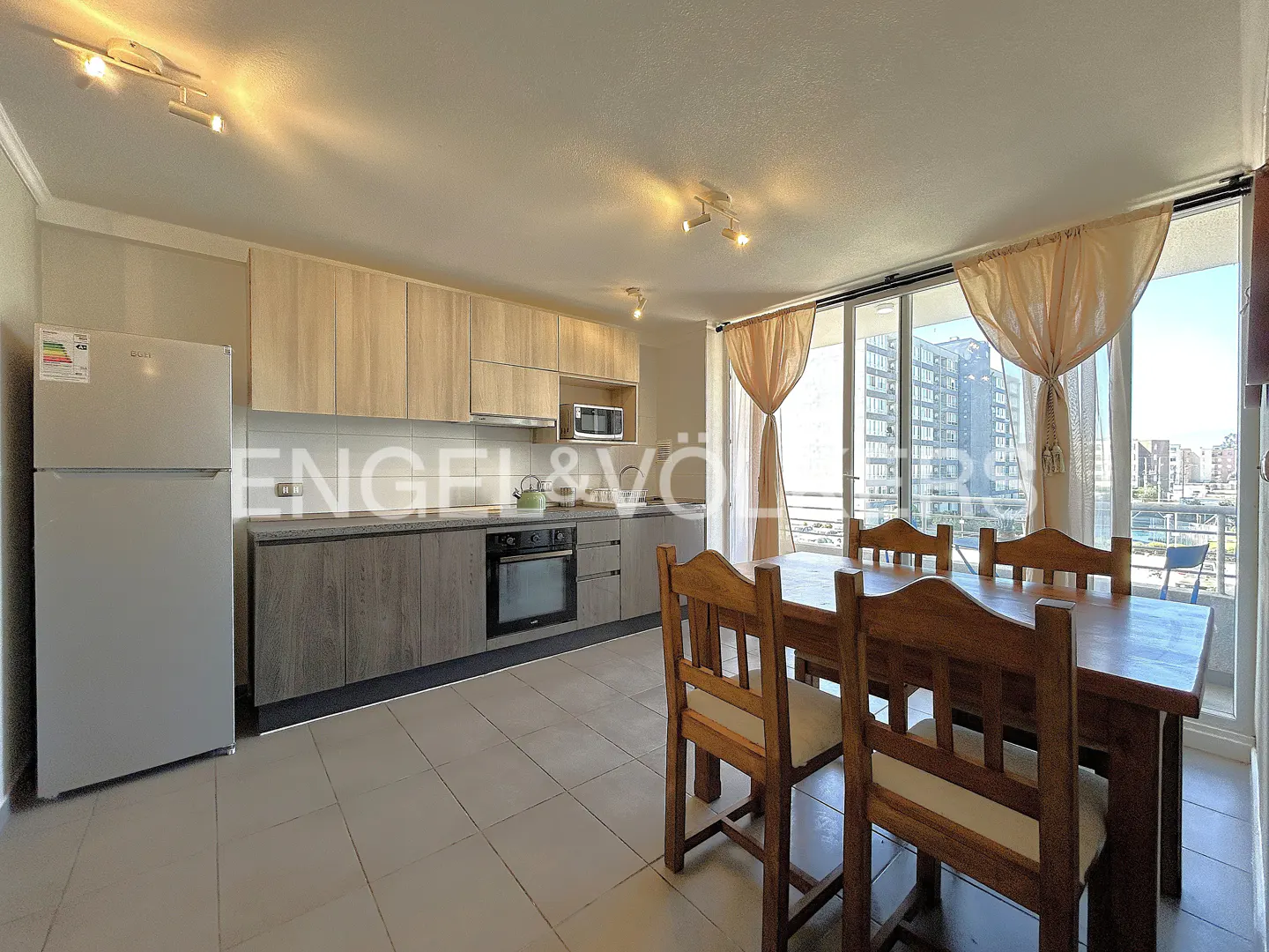 A bright kitchen with light wood cabinets, stainless steel appliances, and a dark wood dining table near a window with sheer curtains.