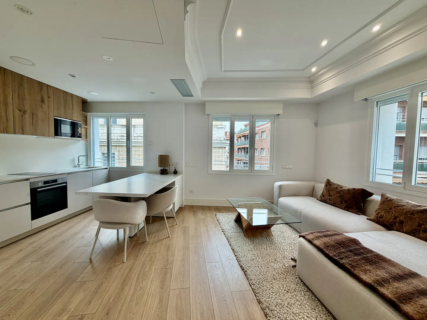 Bright, modern apartment with wood floors. White kitchen cabinets and island with chairs. Beige sofa with brown pillows and glass coffee table.