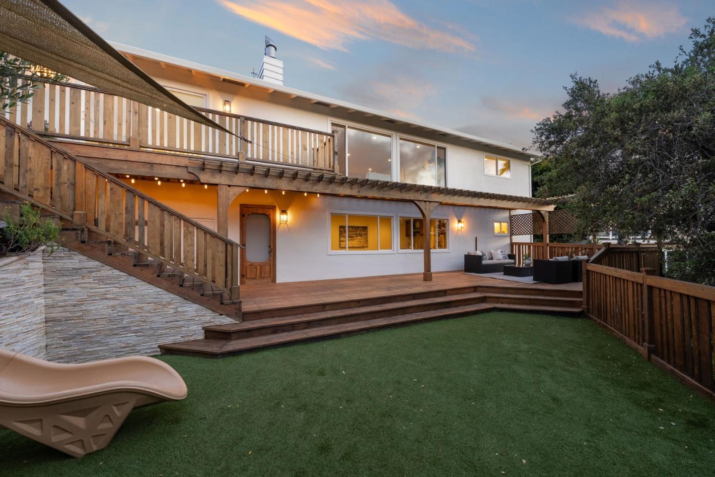 Backyard view of a two-story white house with a wooden deck, stairs, and a slide on green grass.