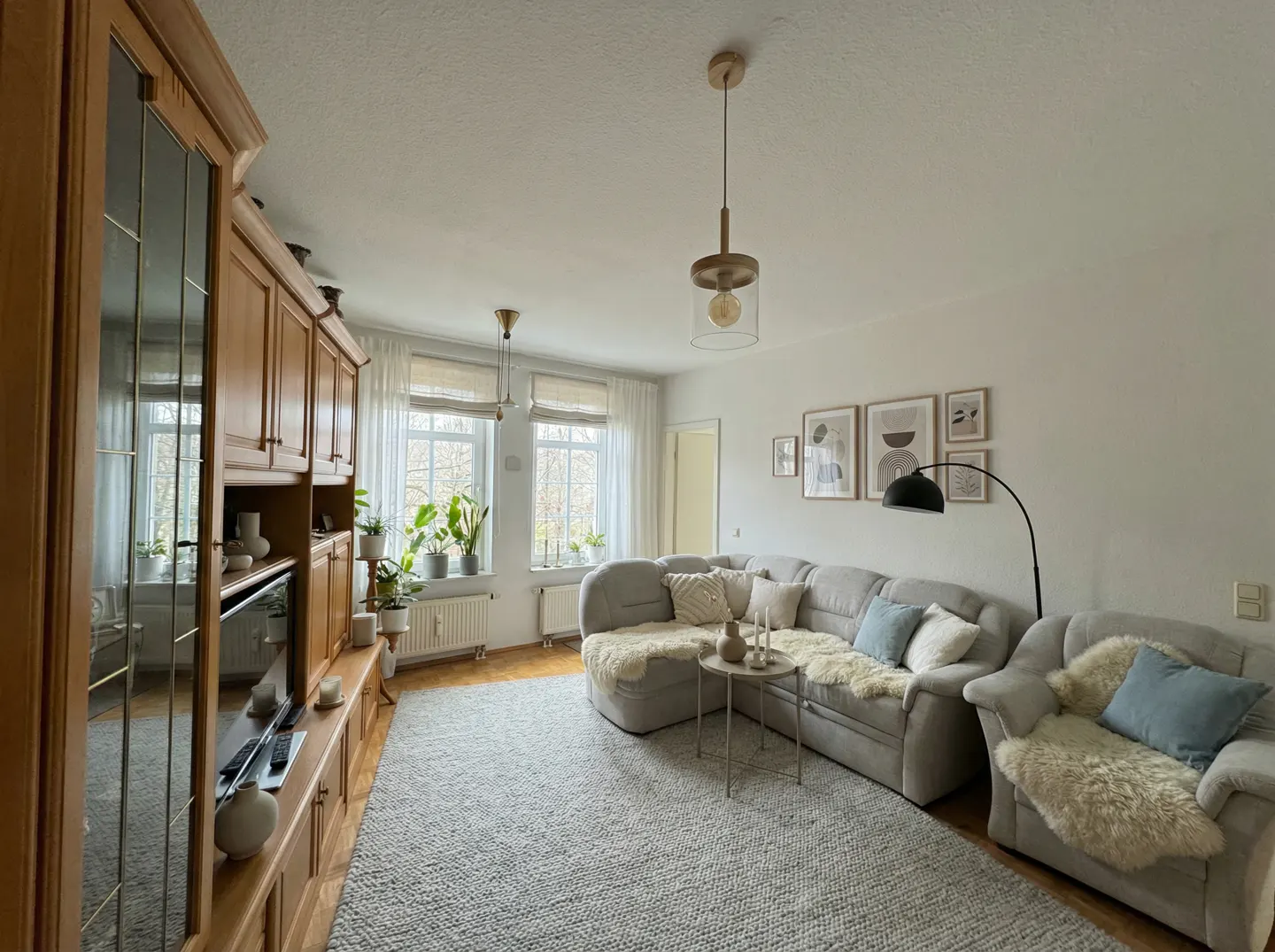 Living room with a gray sectional sofa, a wooden cabinet, and a gray rug. Windows with white curtains and plants are in the background.