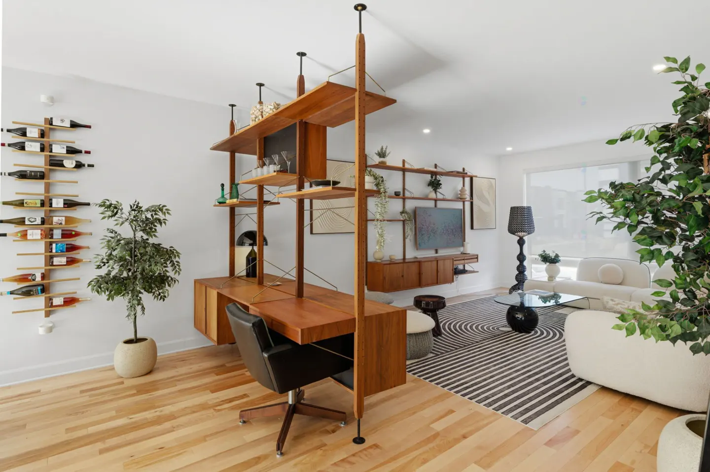 Bright living room with wood floors, a wooden shelving unit with a desk, and a black office chair. A wine rack is on the wall. A white sofa is in the background.