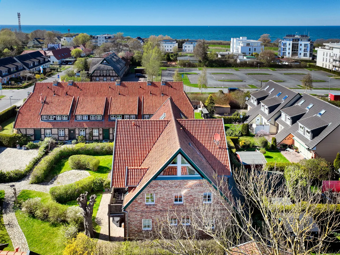Aerial view of red-roofed houses near the sea, with green lawns and trees. Blue sky.