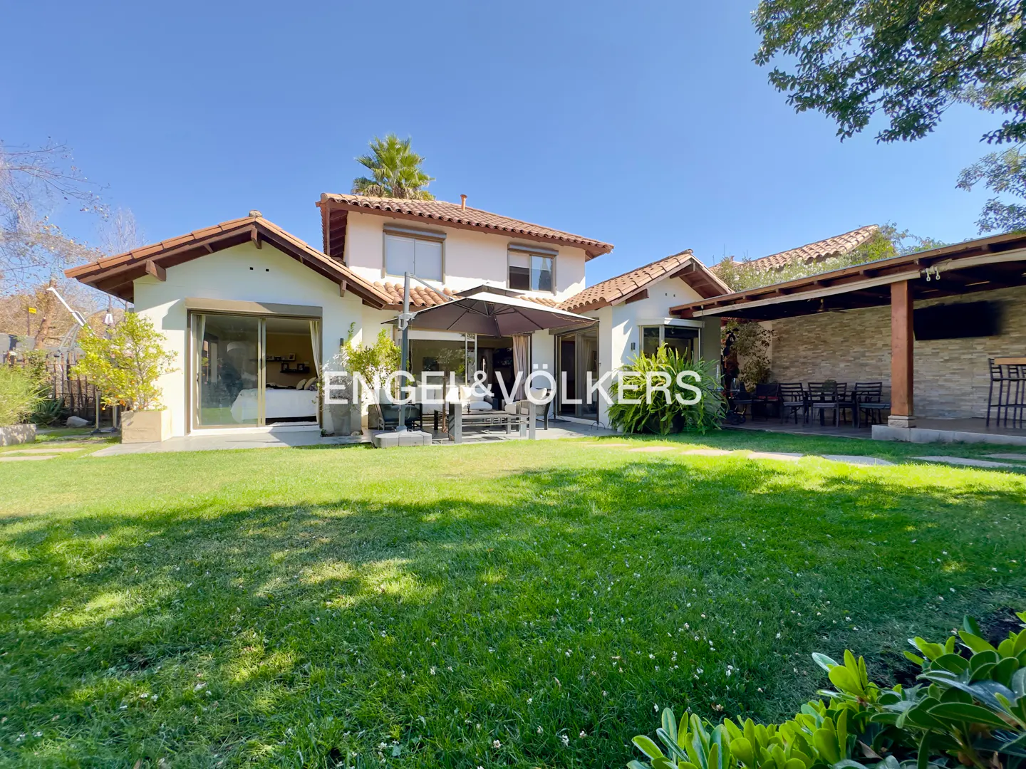Backyard view of a two-story house with a red tile roof, a patio with an umbrella, and a large green lawn.