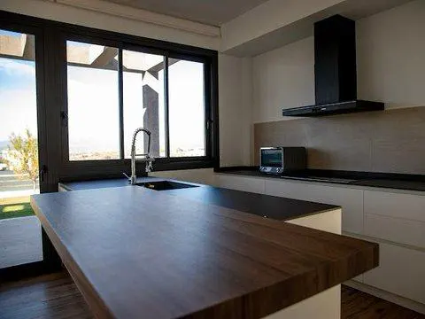 A modern kitchen with dark wood island, black sink, and large window. White cabinets and a black range hood complete the space.
