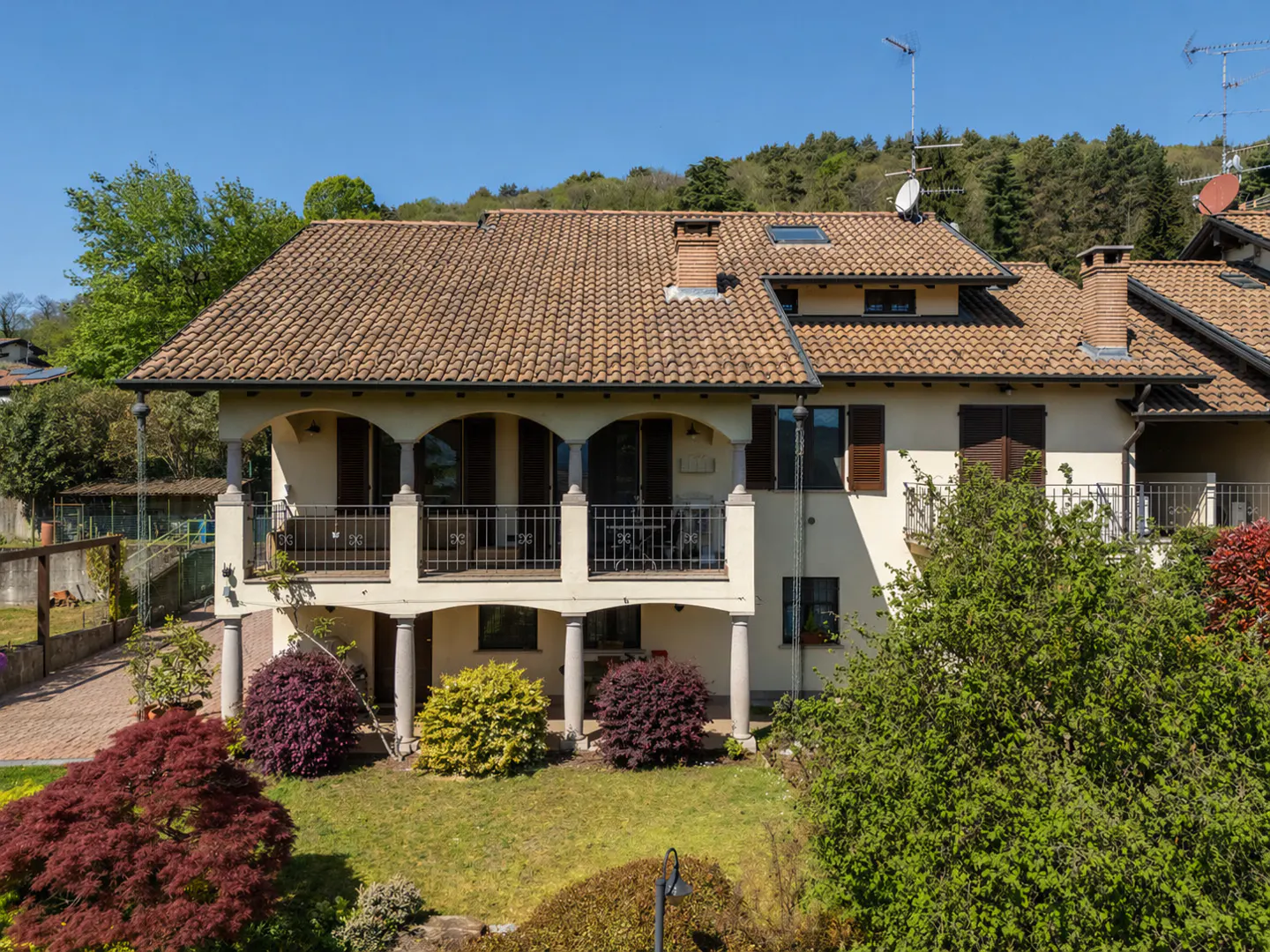 Two-story house with a tan tile roof, cream-colored walls, and a balcony with pillars, surrounded by green trees and bushes.