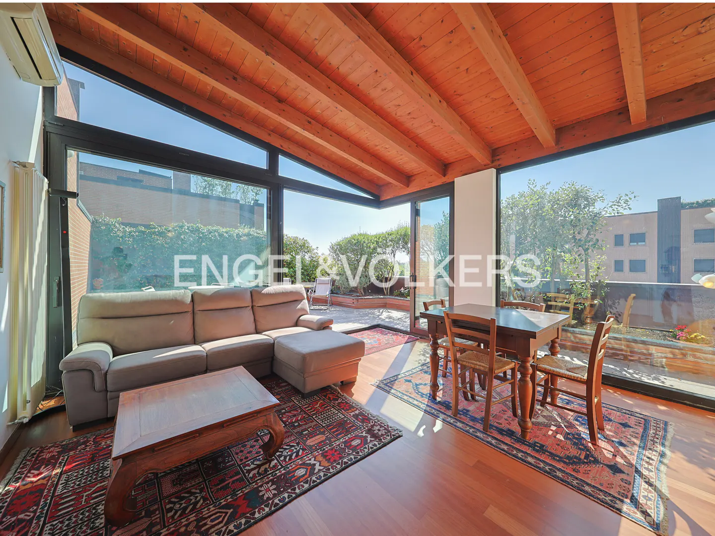 Sunlit living room with wood-beamed ceiling, beige sectional sofa, wooden dining set, and large windows opening to a patio.