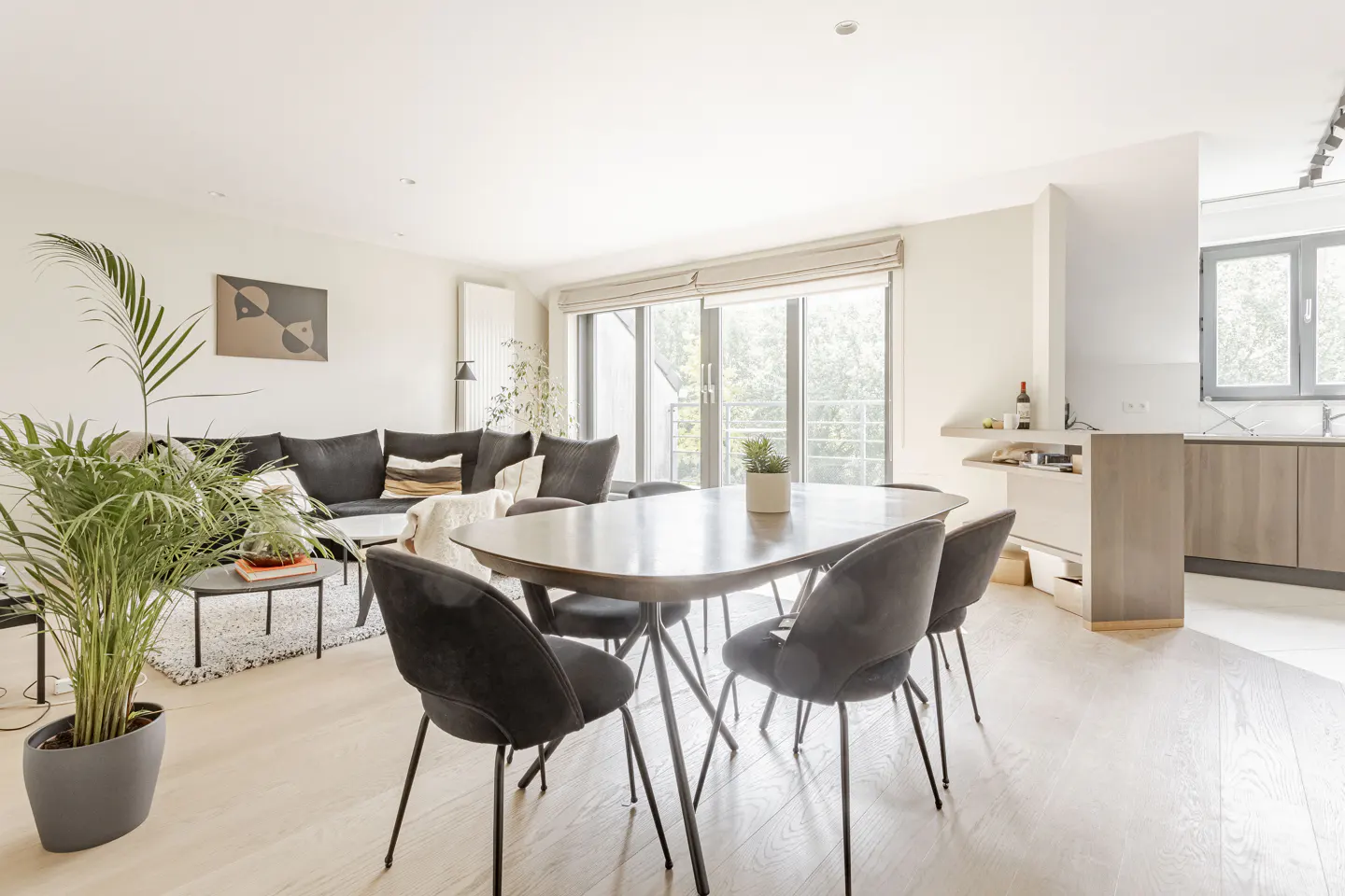 Bright, open-plan living space with a dark wood dining table, black chairs, and a dark gray sectional sofa. Large windows provide natural light.
