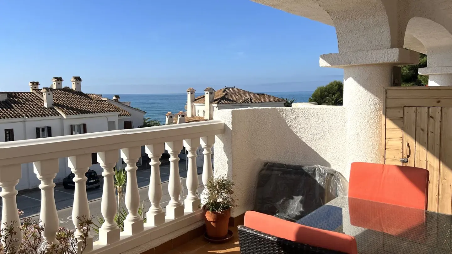 Balcony view of ocean and white buildings. Orange cushions on wicker chairs surround a glass table. White balustrade.