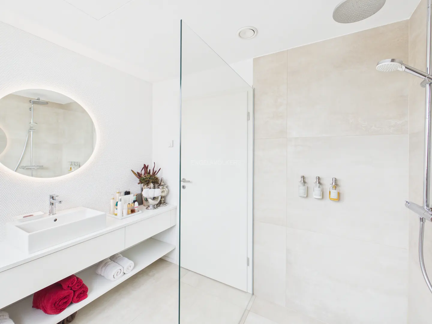 Bright, modern bathroom with white vanity, sink, and oval mirror. Red towels are on the shelf. Glass shower door and tiled shower area.