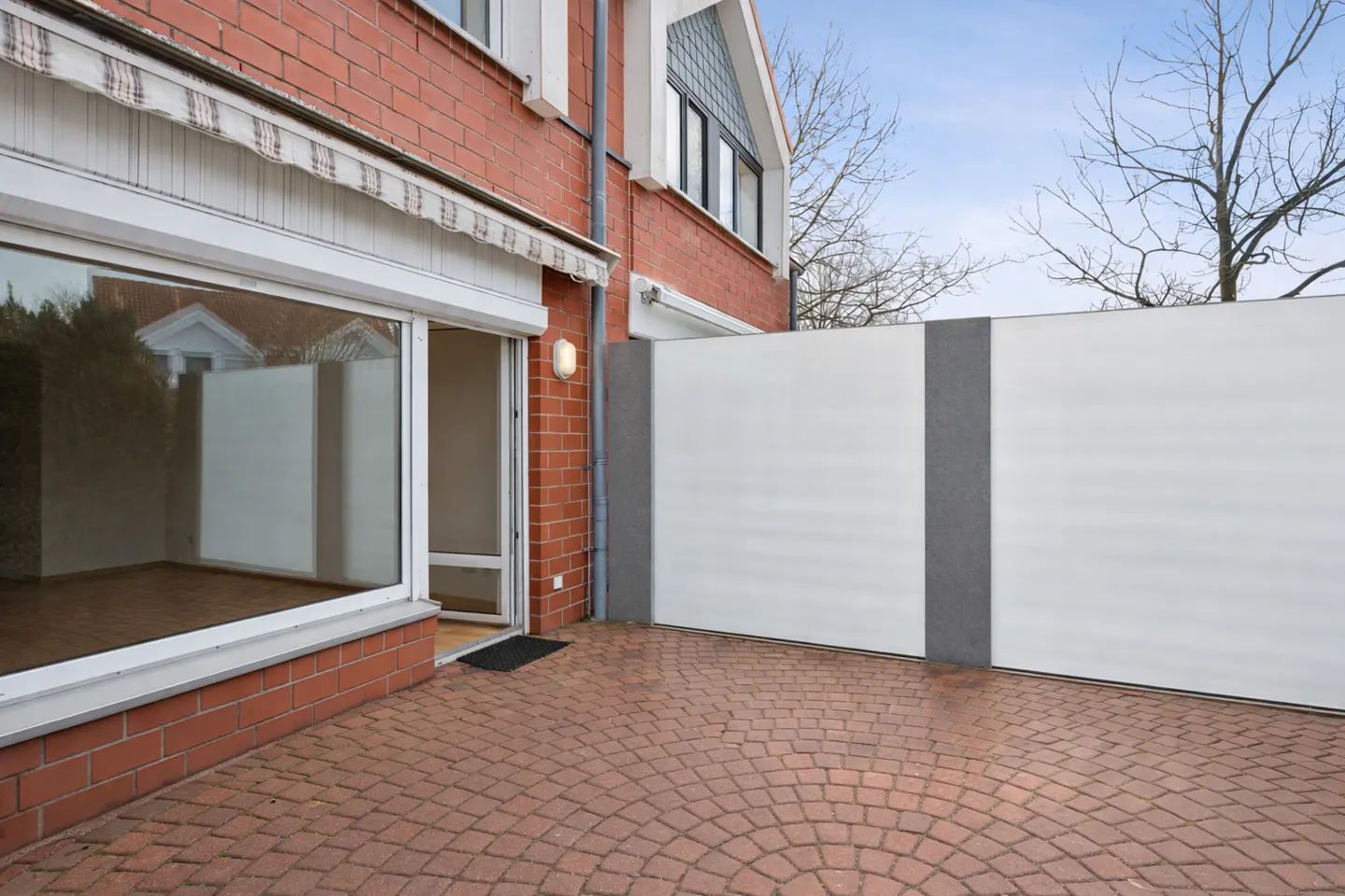 Brick patio with a red brick house, large window, open door, and white privacy fence.