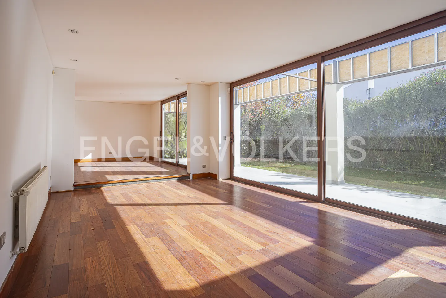 Bright, empty living room with hardwood floors and large windows overlooking a green yard. White walls and radiator. Engel & Volkers logo visible.