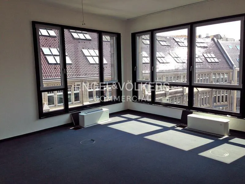 Empty office space with blue carpet, white walls, and large windows. Building with red roofs visible through the windows.