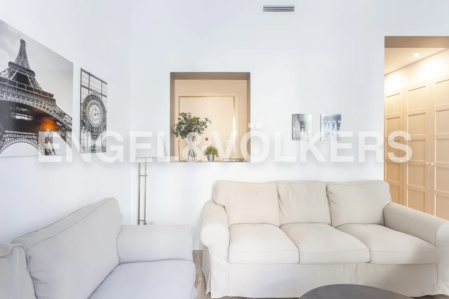 Living room with white walls, a beige sofa, a wall niche with plants, and black-and-white Eiffel Tower wall art.