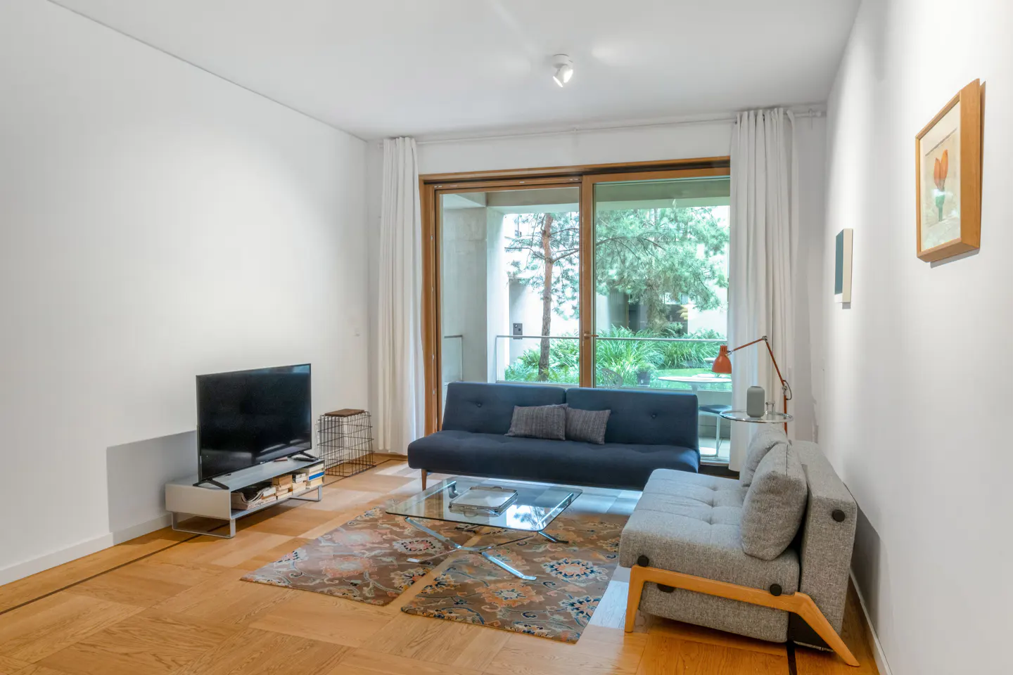 Living room with a blue sofa, gray chair, glass table, TV, and a view of a green garden through sliding glass doors.