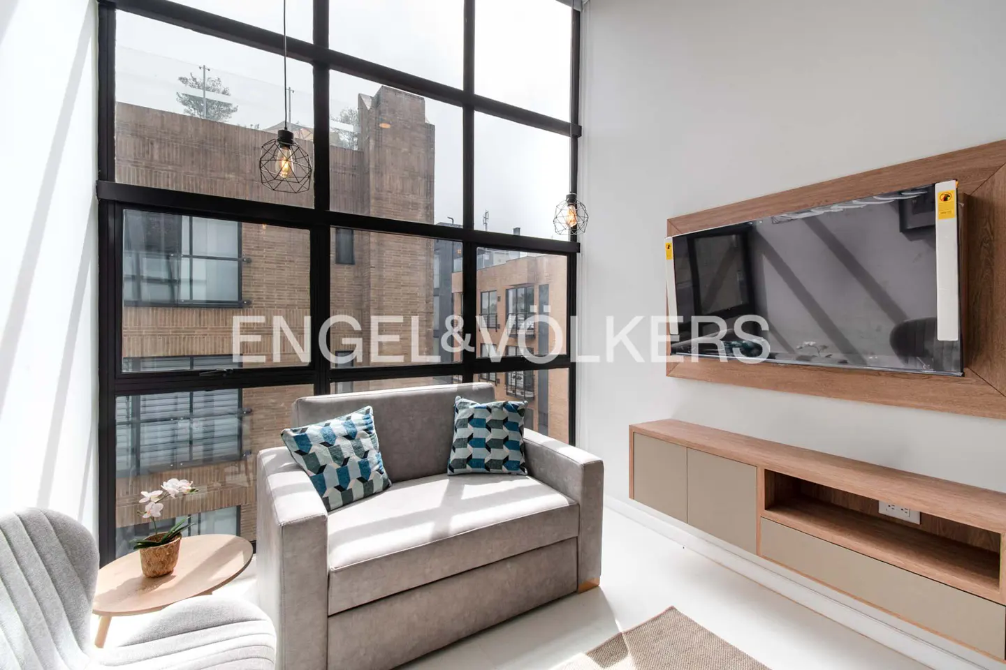 Living room with a gray sofa, blue pillows, and a large window with a city view. A TV is mounted on the wall above a beige cabinet.