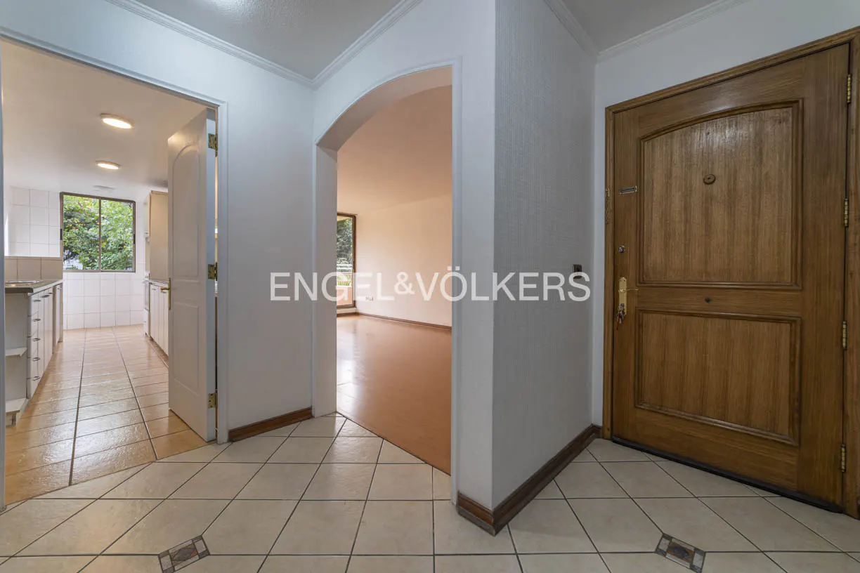A home's entryway with a wooden front door, tiled floor, and arched doorway leading to a living room. A kitchen is visible through another doorway.