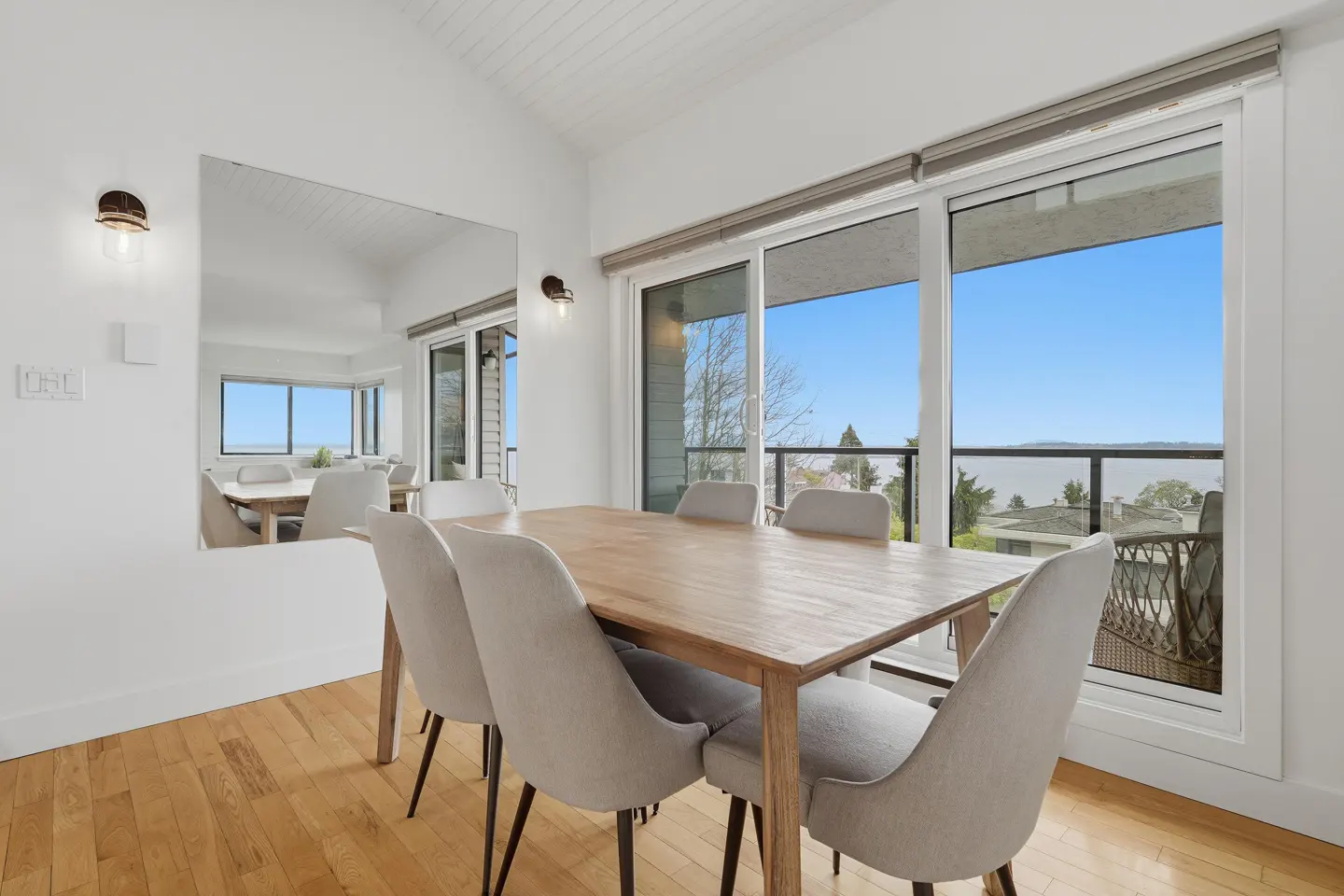 Bright dining room with wood table, six gray chairs, and a large window with a view of the water. A mirror reflects the room.