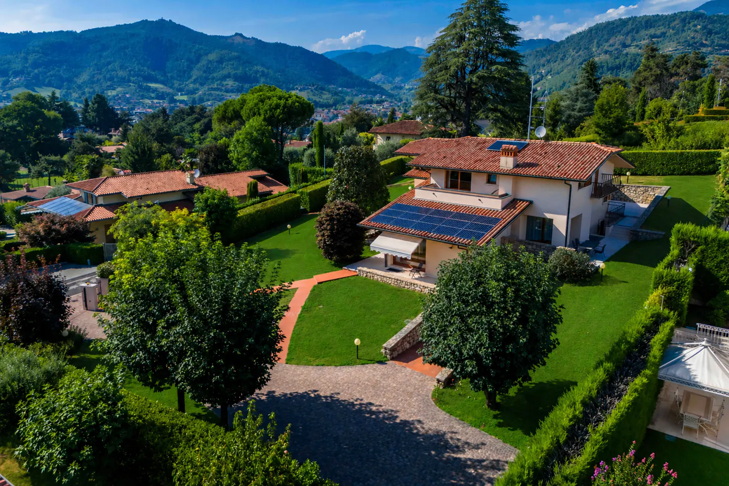 Aerial view of a cream-colored house with a red tile roof and solar panels, surrounded by green lawns, trees, and mountains in the background.