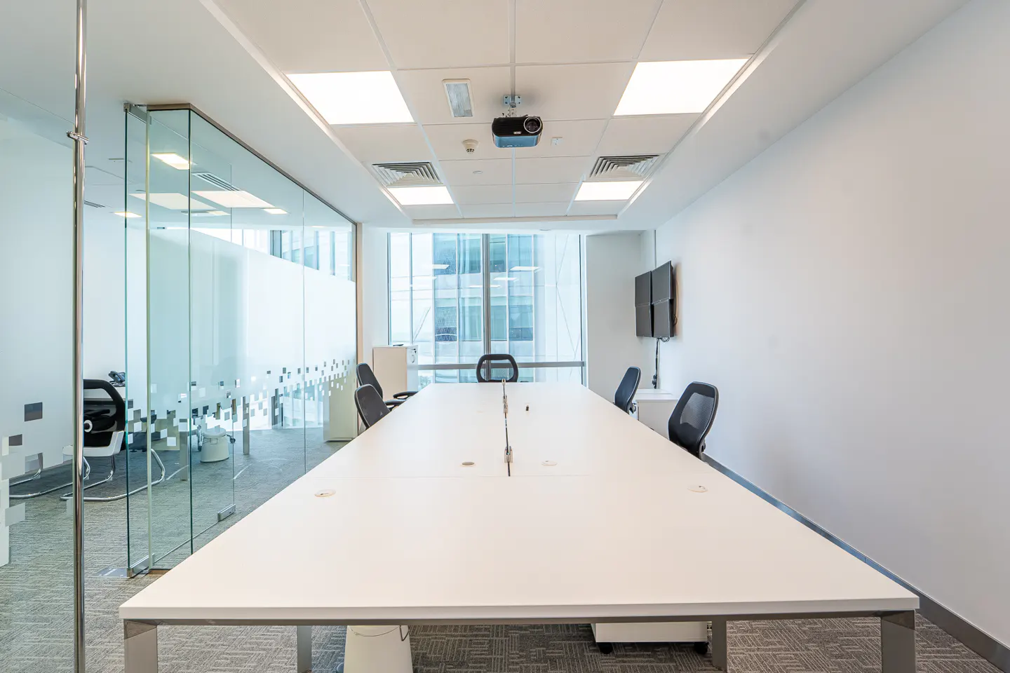 Bright, modern office meeting room with a long white table, black chairs, glass walls, and a projector on the ceiling.
