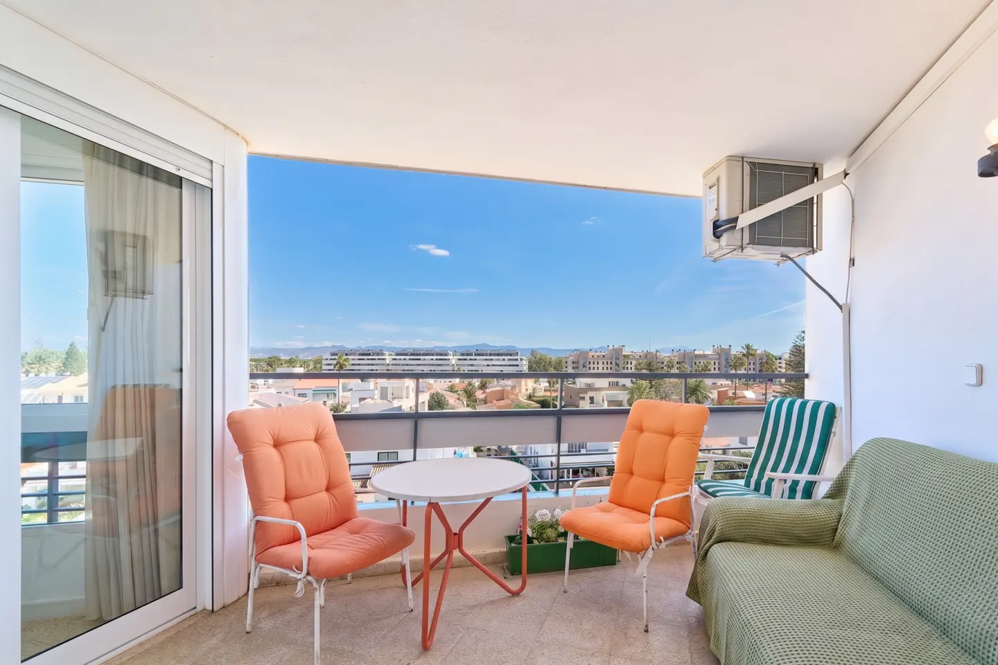 Balcony with orange chairs, a white table, and a green sofa overlooking a city under a blue sky.