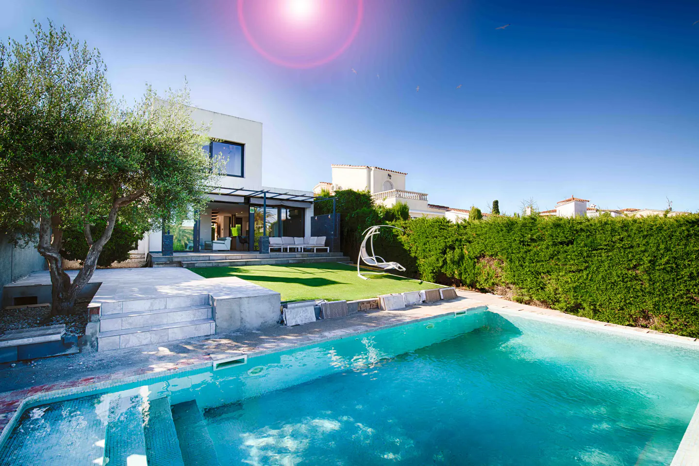 A modern home with a pool, green lawn, and a white hanging chair under a bright blue sky.