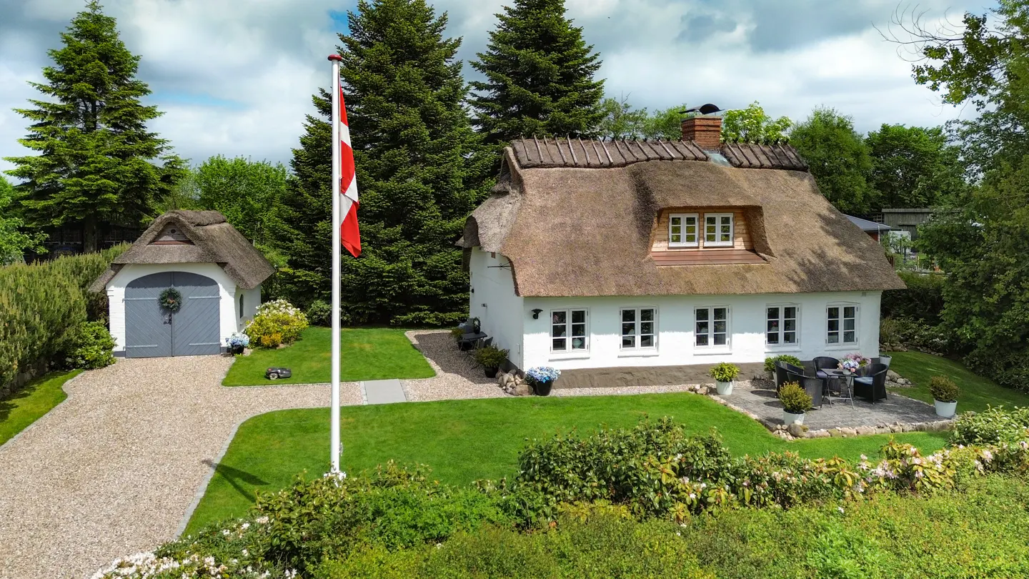 White cottage with a thatched roof, a detached garage, a Danish flag, and a patio set in a lush green garden.