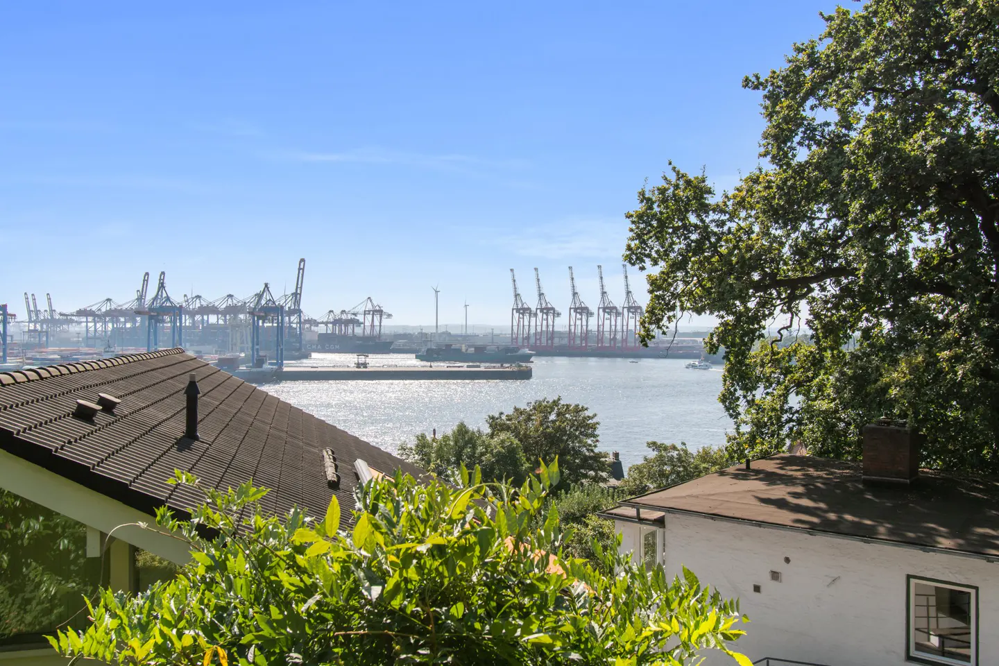 View of a harbor with cranes from a house. The sky is blue and the water is sparkling.