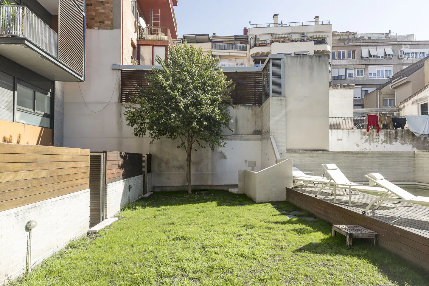 Backyard with green grass, a tree, and white lounge chairs on a wooden deck next to a pool, surrounded by buildings.