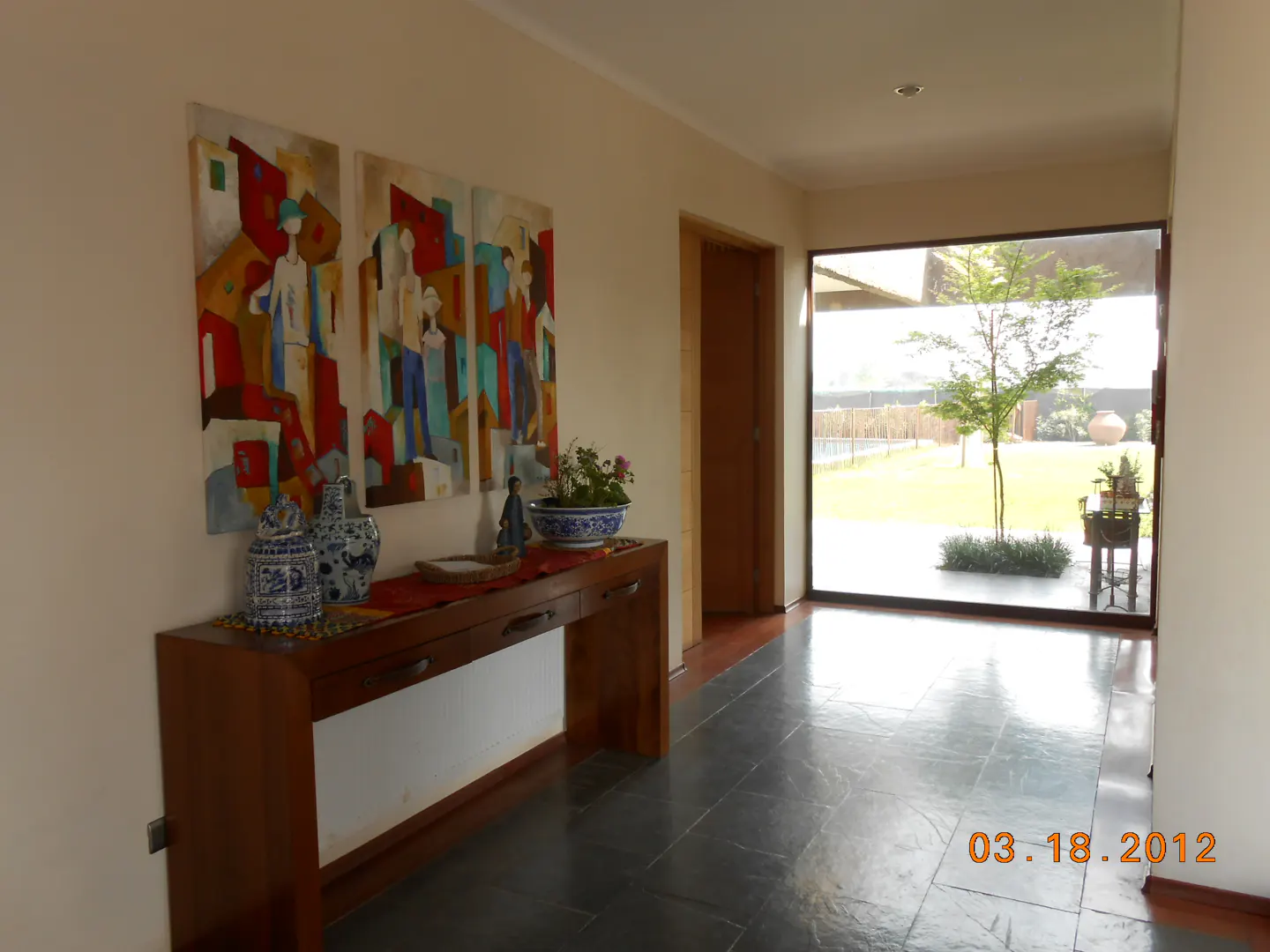Hallway with a wood console table, blue and white vases, and colorful art. A large window shows a green lawn and tree.