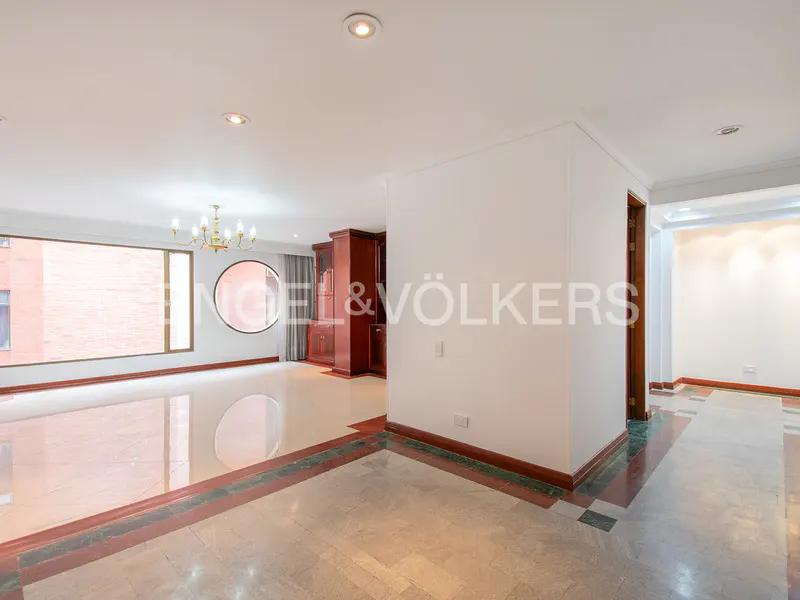 Bright, open living space with marble floors, large windows, and a chandelier. A dark wood cabinet stands against a white wall.