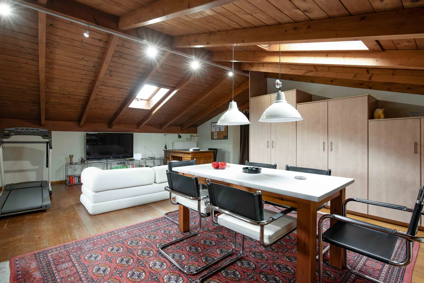 Attic apartment with wood ceiling, skylights, and a red patterned rug. A white table and black chairs sit near a white sofa and treadmill.
