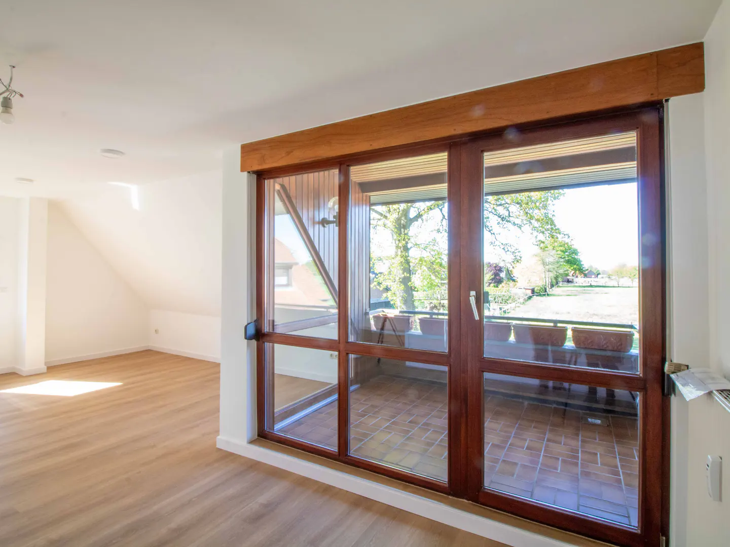 Bright room with light wood floors and large wooden-framed glass doors opening to a tiled balcony overlooking a garden.