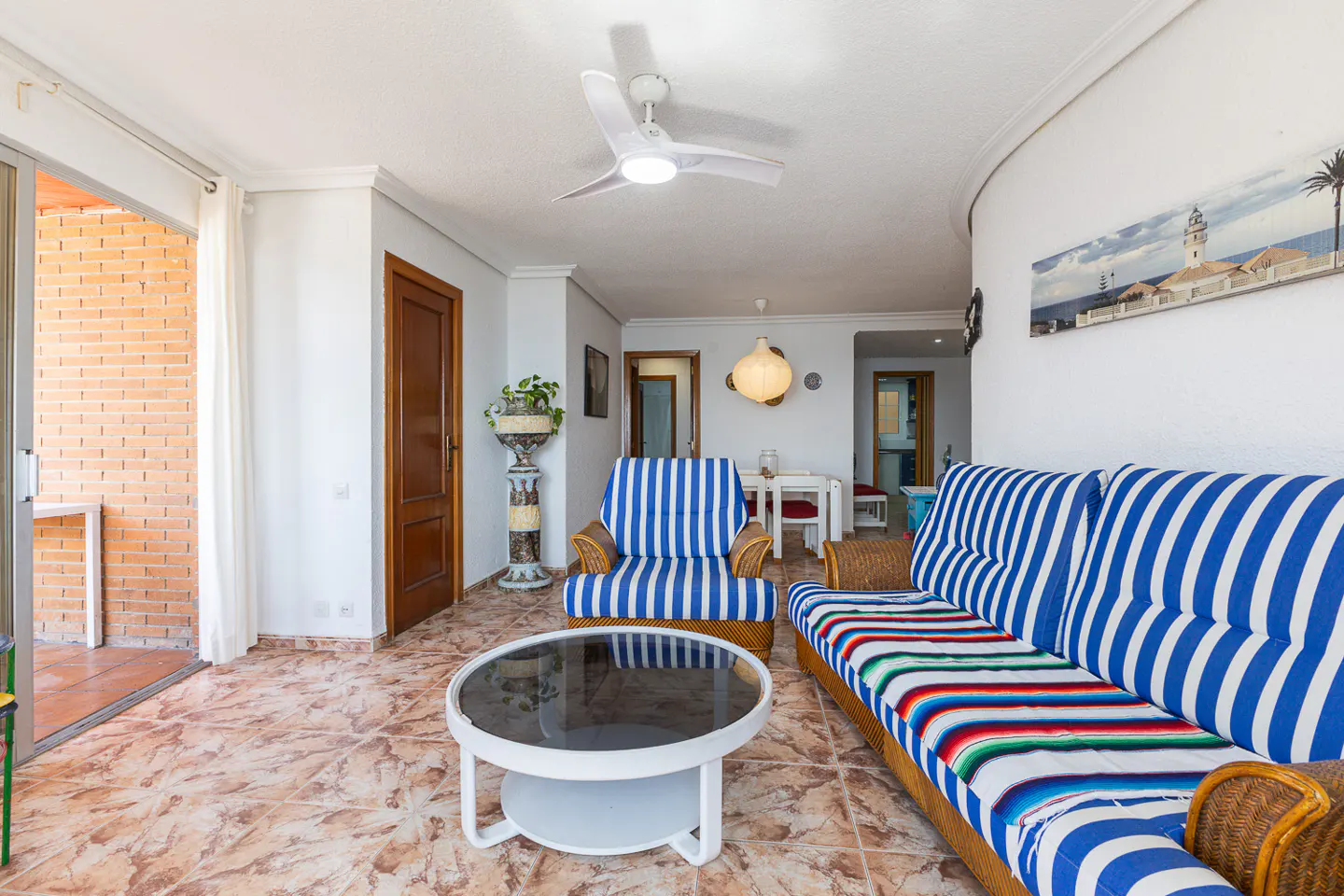 Bright living room with blue and white striped sofa and chair, round glass table, and balcony with brick wall.