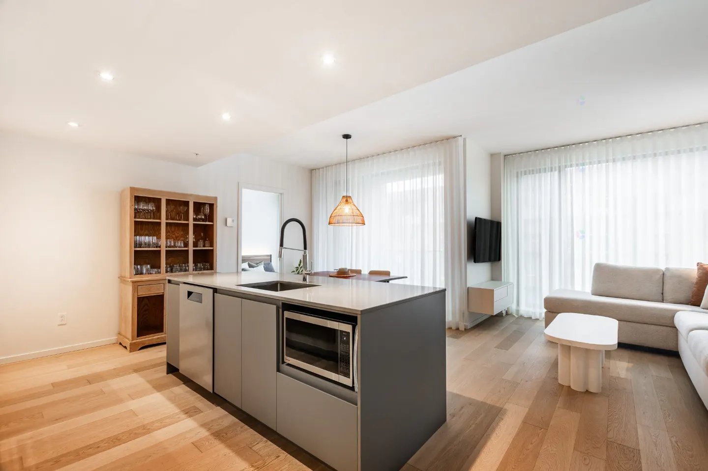 Bright, modern open-concept living space with a gray kitchen island, wood floors, and a beige sofa near sheer white curtains.