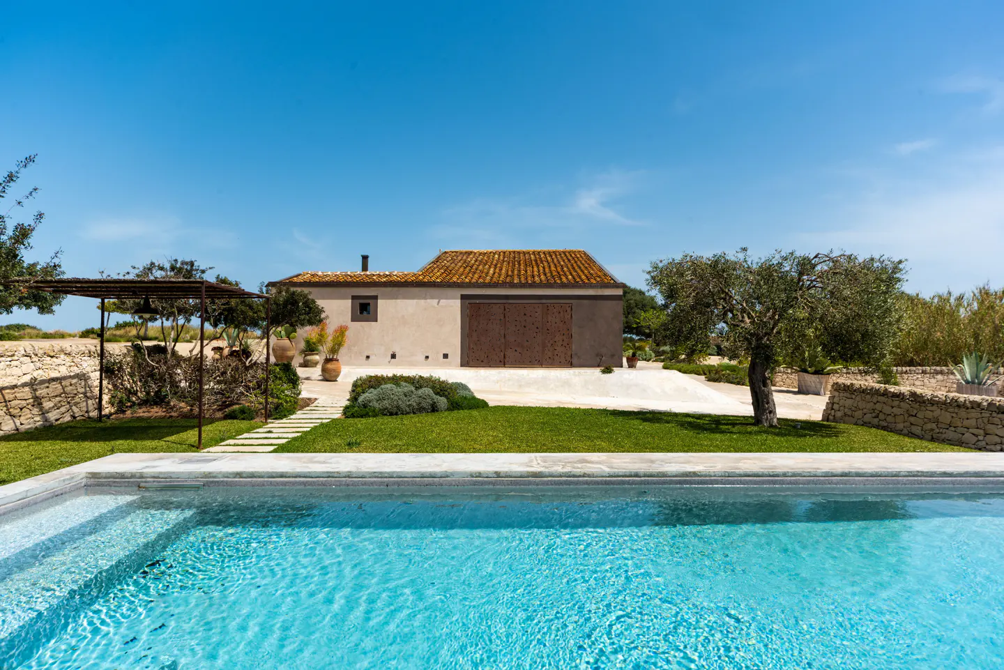 A sunny view of a turquoise pool, green lawn, and a beige building with a brown tile roof under a clear blue sky.