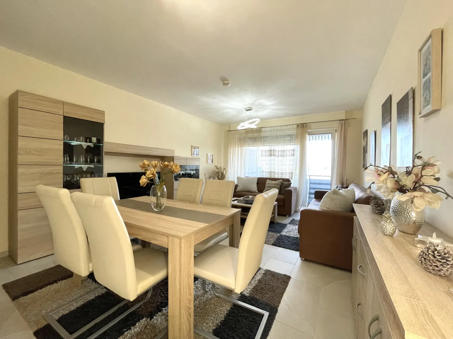 A bright living and dining area with light wood furniture, cream chairs, and brown sofas. A rug with brown and black patterns is on the floor.