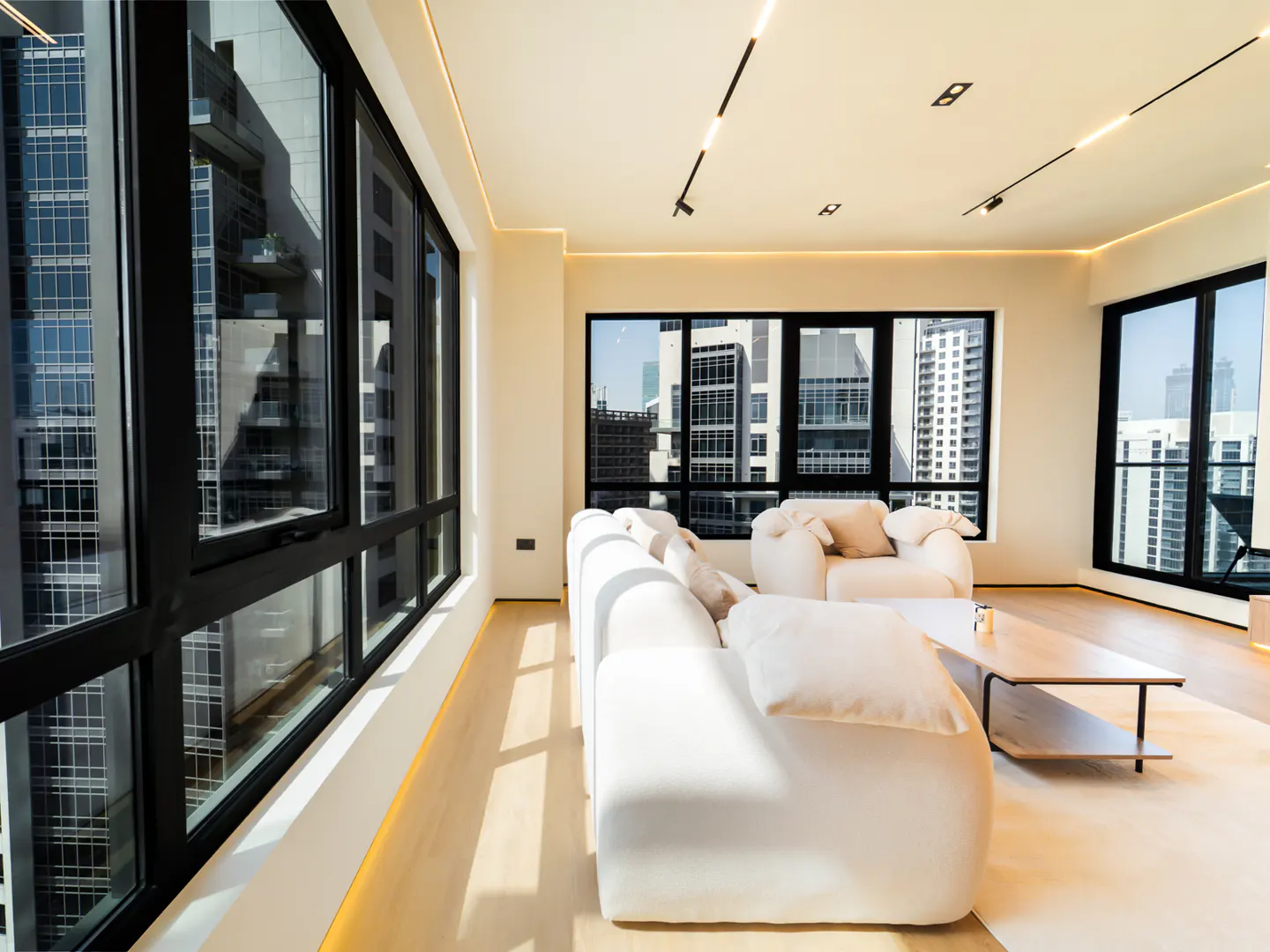 Bright, modern living room with white sofa and chair. Large windows offer city views. Light wood floors and minimalist decor.