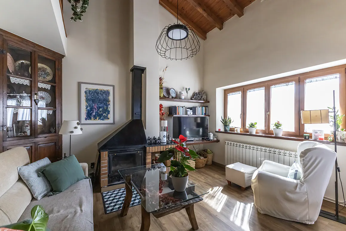 Bright living room with a fireplace, wood floors, and large windows. A sofa, armchair, and glass coffee table furnish the space.