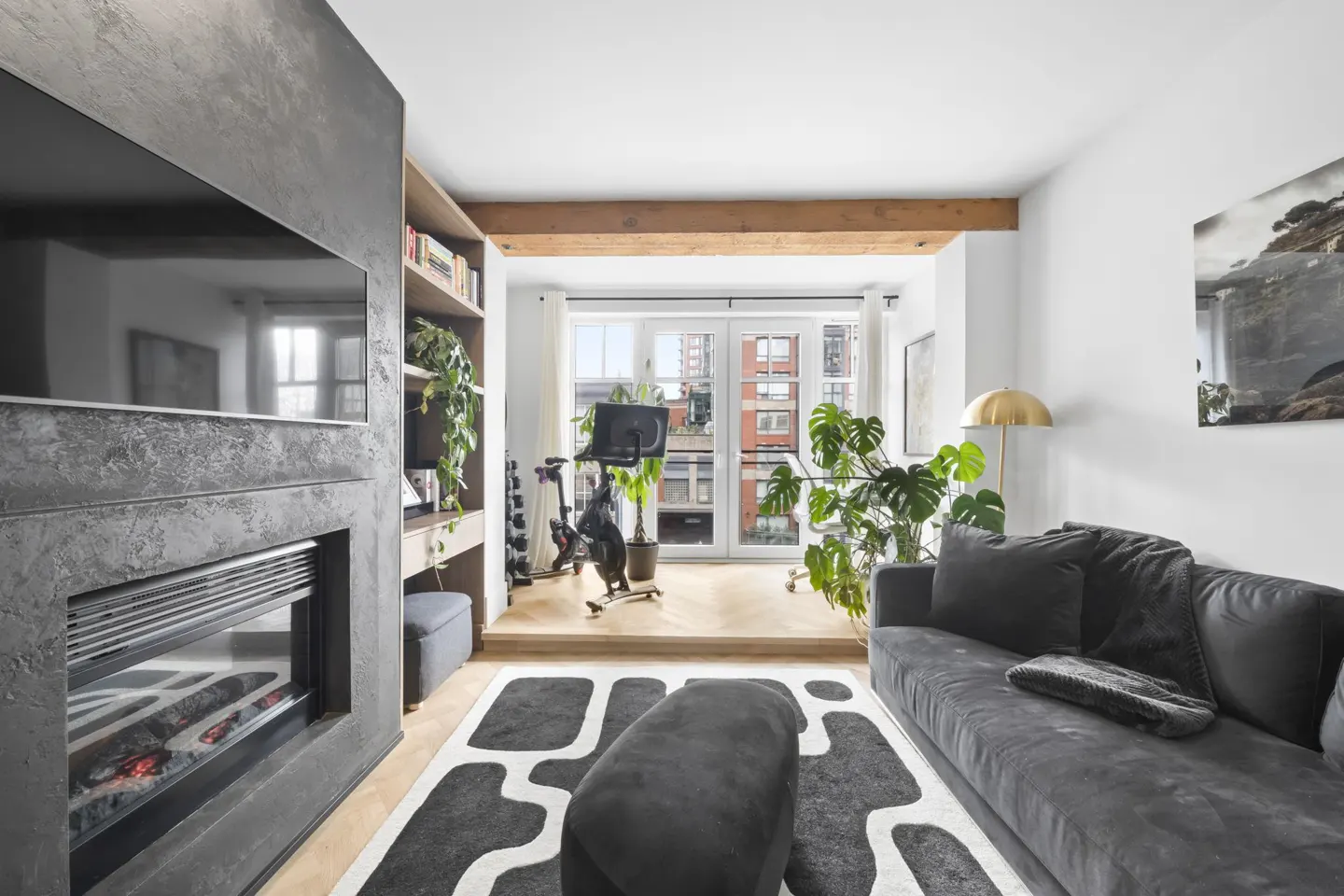Living room with a gray sofa, black and white rug, fireplace, TV, and exercise bike near the balcony.