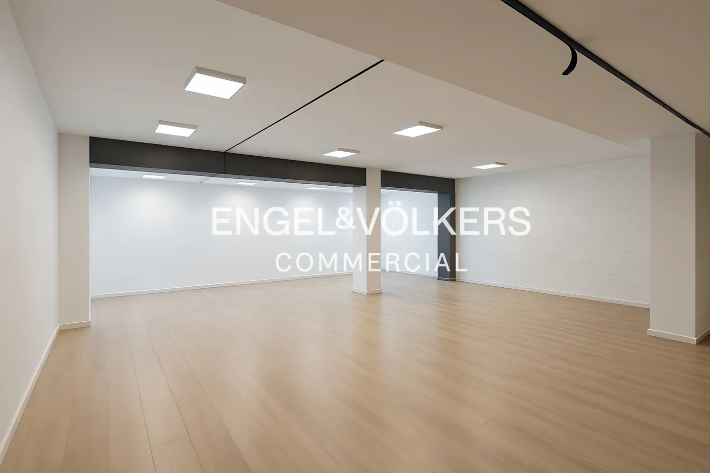 Empty commercial space with light wood floors, white walls, and square ceiling lights. "Engel & Völkers Commercial" is superimposed on the image.