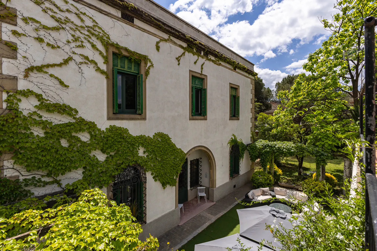 Exterior view of a white building with green shutters and ivy, overlooking a garden with white umbrellas and seating.