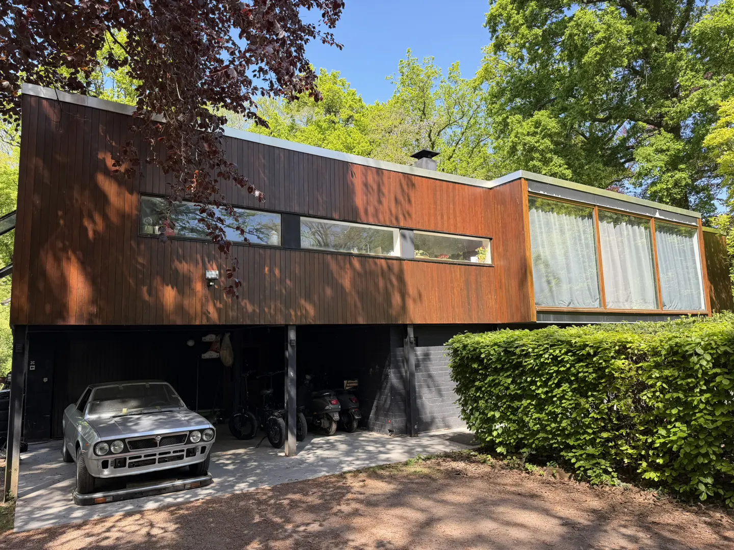 Modern wood-paneled house with a car parked underneath. Green trees surround the property.