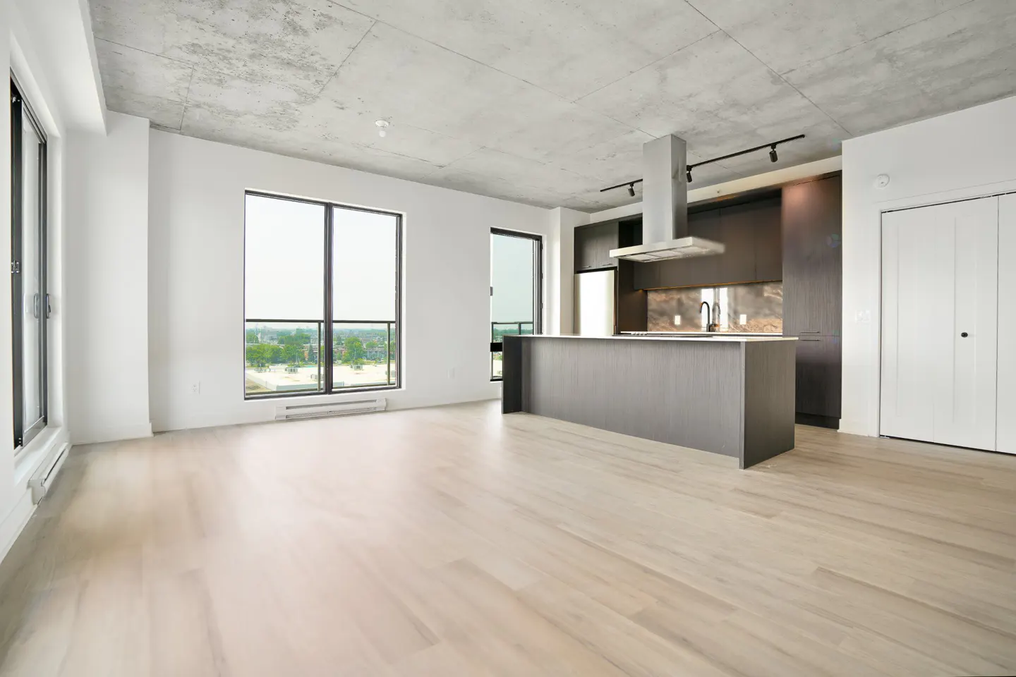 Modern open-concept kitchen with dark cabinetry, a central island, and light wood floors under a raw concrete ceiling.
