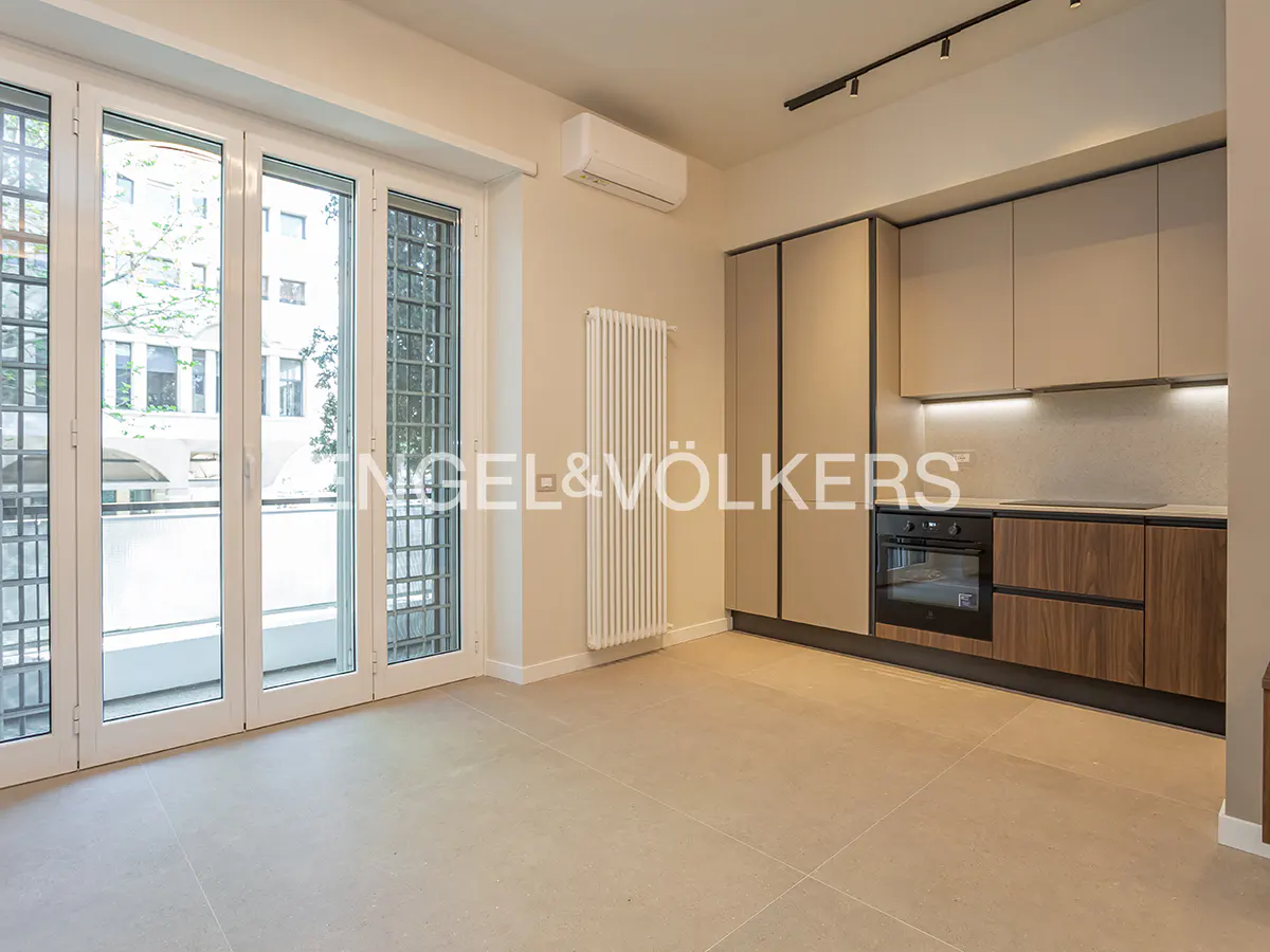 A bright, empty apartment with a modern kitchen and large windows. The kitchen has light gray cabinets and dark wood lower cabinets. The floor is light beige tile.