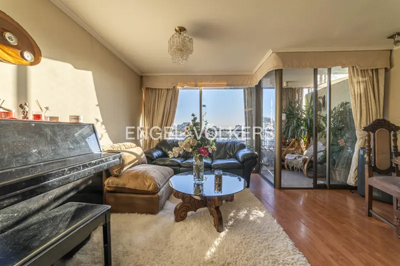 A living room with a piano, black leather sofa, glass table, and a balcony with plants. The room has a white rug and hardwood floors.