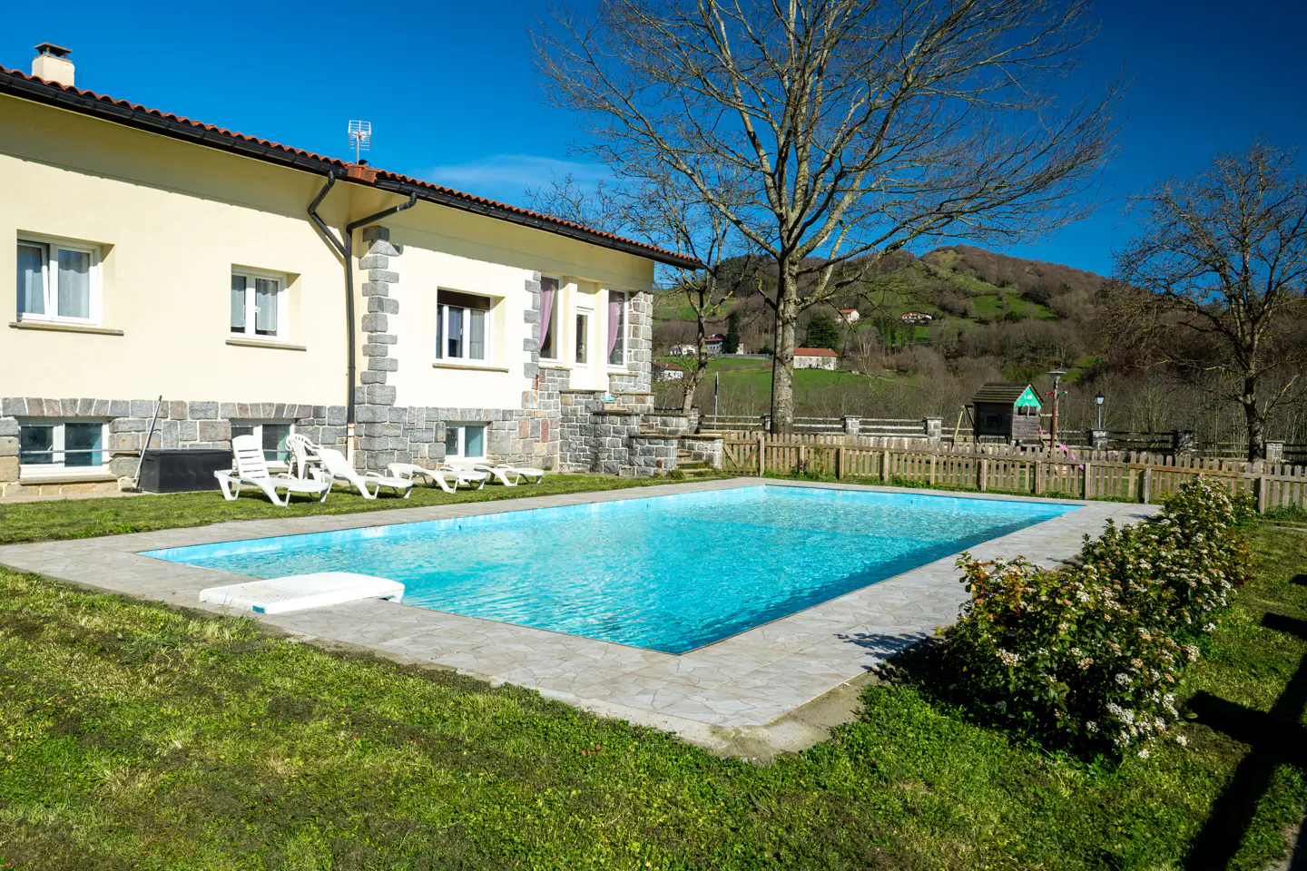 A cream-colored house with stone accents sits beside a bright blue swimming pool, set against a backdrop of green hills.