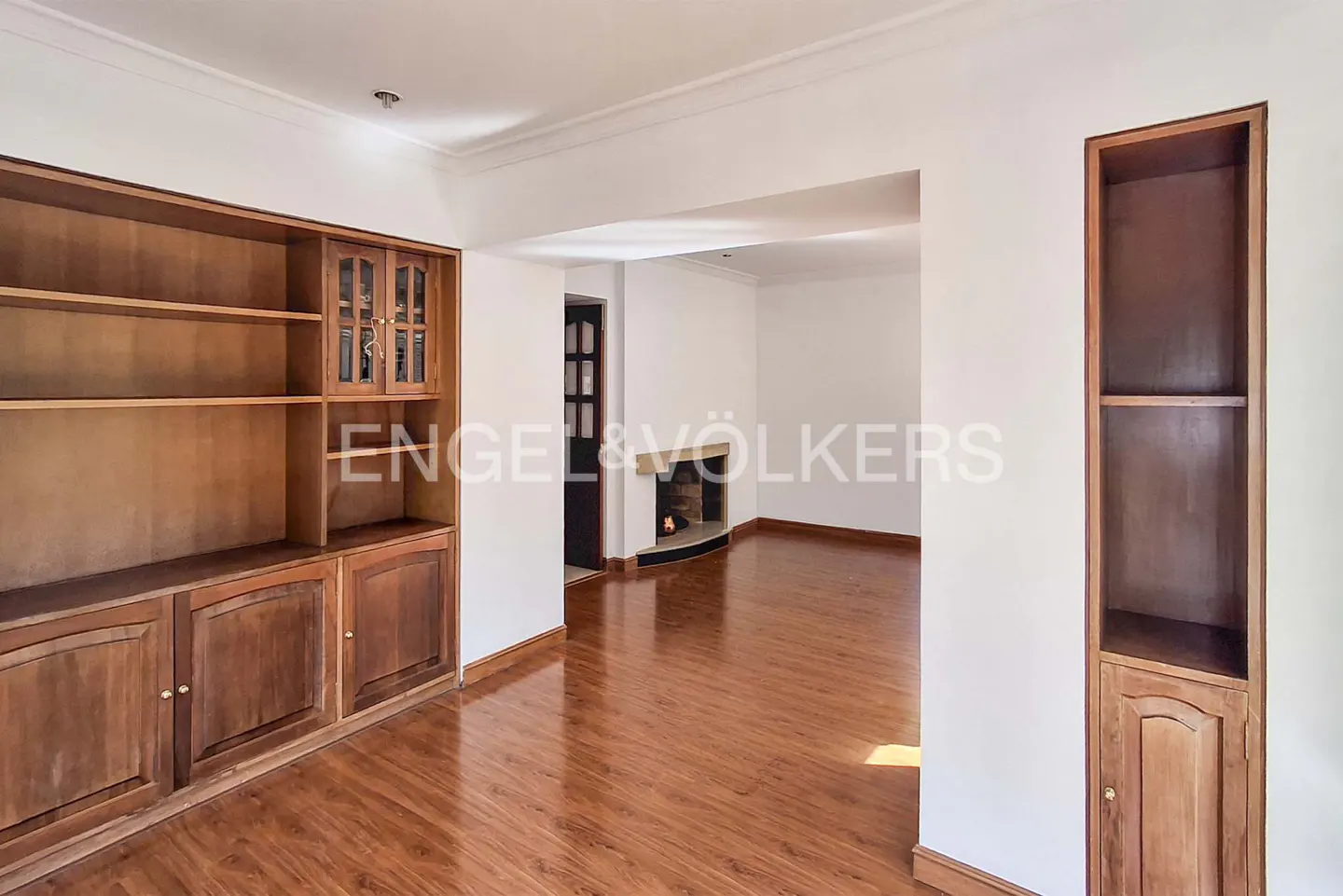 Living room with wood floors, built-in wooden shelving units, and a distant fireplace in a white-walled interior.
